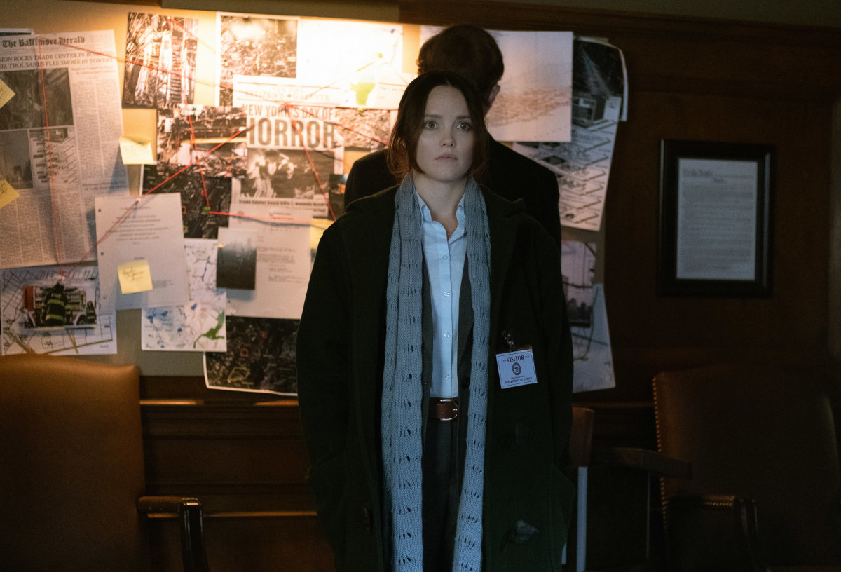 Clarice looking at the camera with a bulletin board of newspaper clippings behind her 