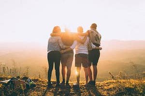 Four young adults facing away from the camera are standing atop the crest of a hill, backlit by the setting sun, which shines a flare between their heads.