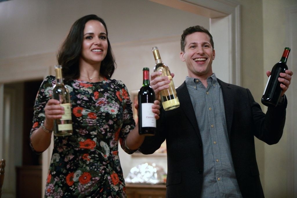 Melissa Fumero and Andy Samberg holding up bottles of wine in a scene from BROOKLYN NINE-NINE