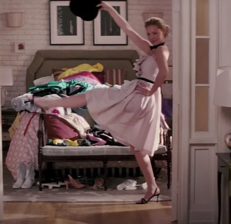 Jane high-kicks in front of a bed wearing a pink knee-length dress and holding a top hat