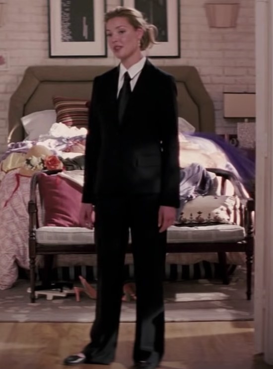 Jane wears a suit and stands in front of a bed