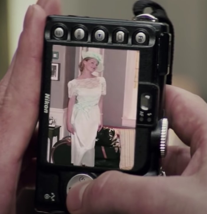 A shot of a digital camera with a picture of Jane on the screen wearing awhite dress with a lacy top half and a mint green ribbon and matching hat