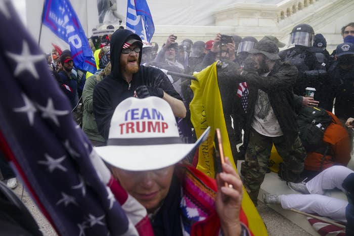 Police and rioters outside the Capitol clash; in the foreground, a person wears a cowboy hat that reads "team Trump"