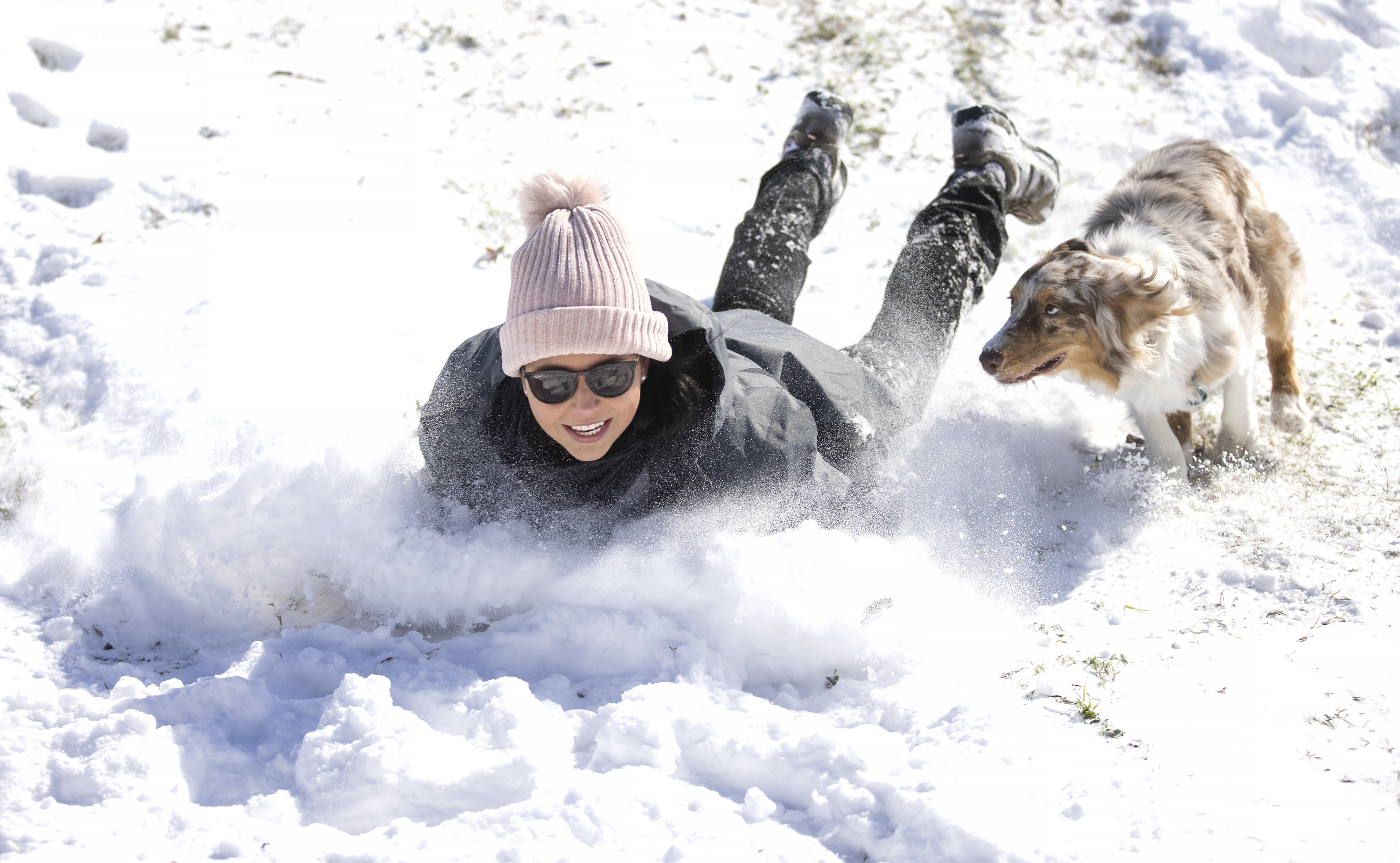 A woman sleds down a hill on her stomach chased by her dog