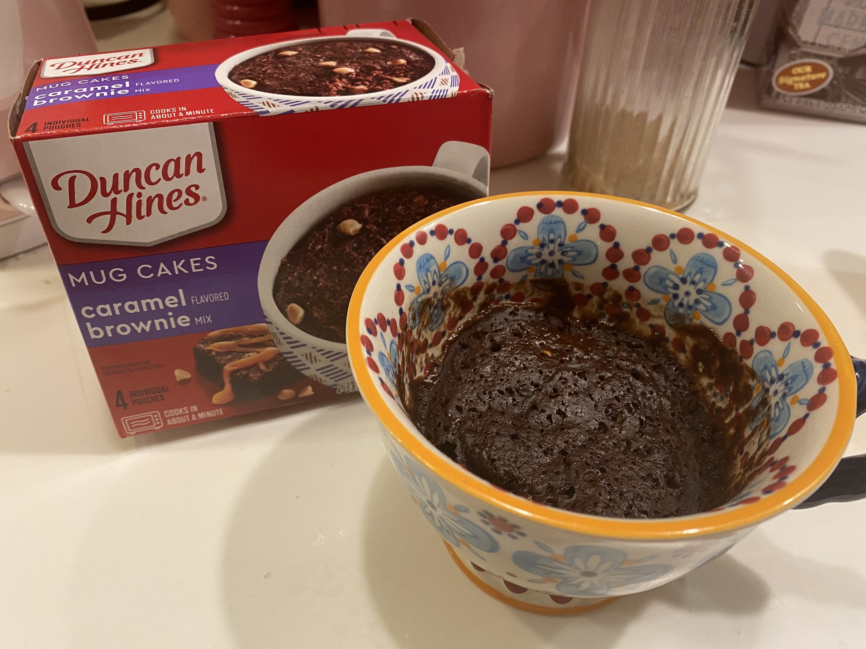 A chocolate caramel brownie cake in a mug next to a box of cake 