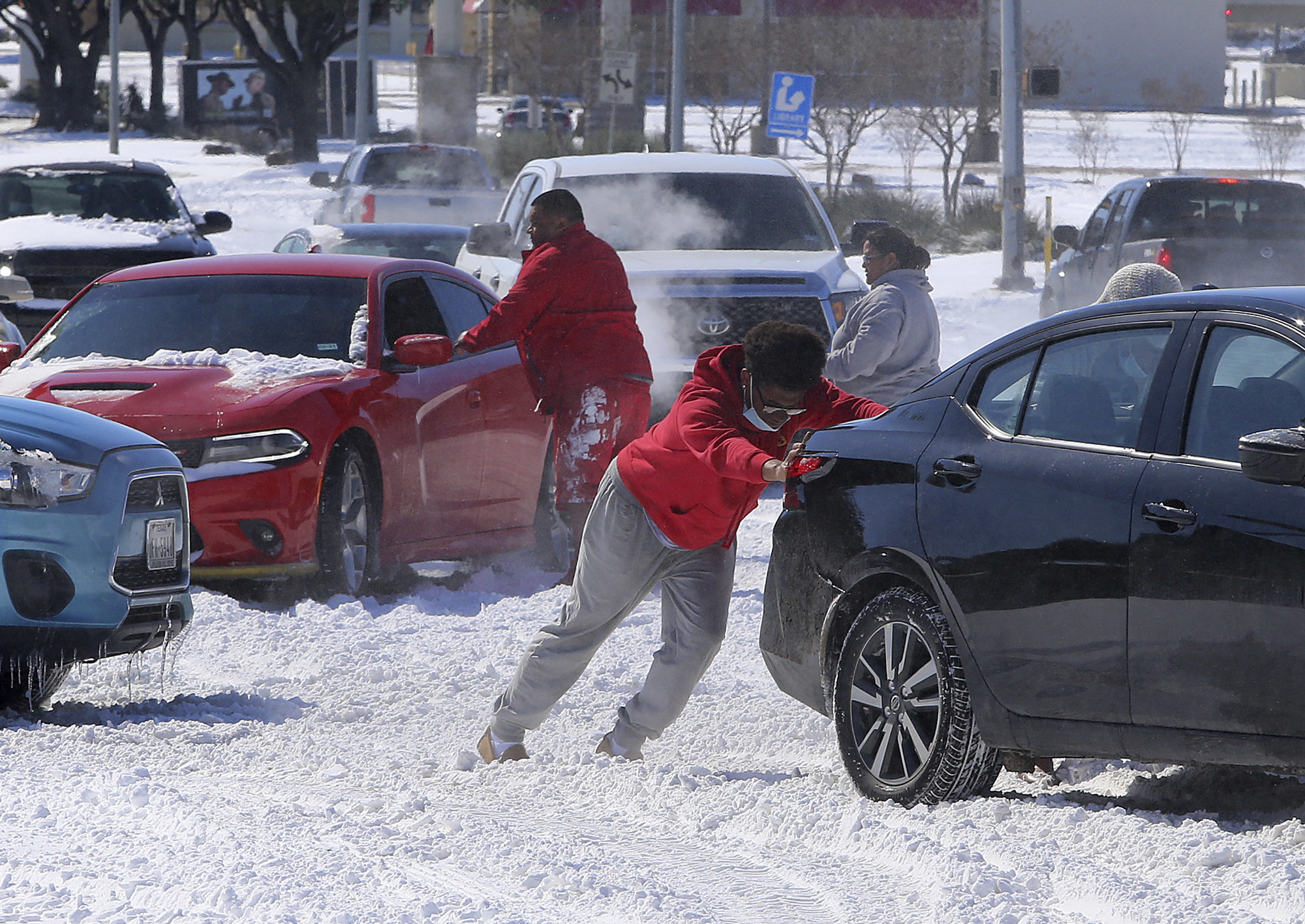 People at a crowded snowy intersection try to push their cars out of harm&#x27;s way 