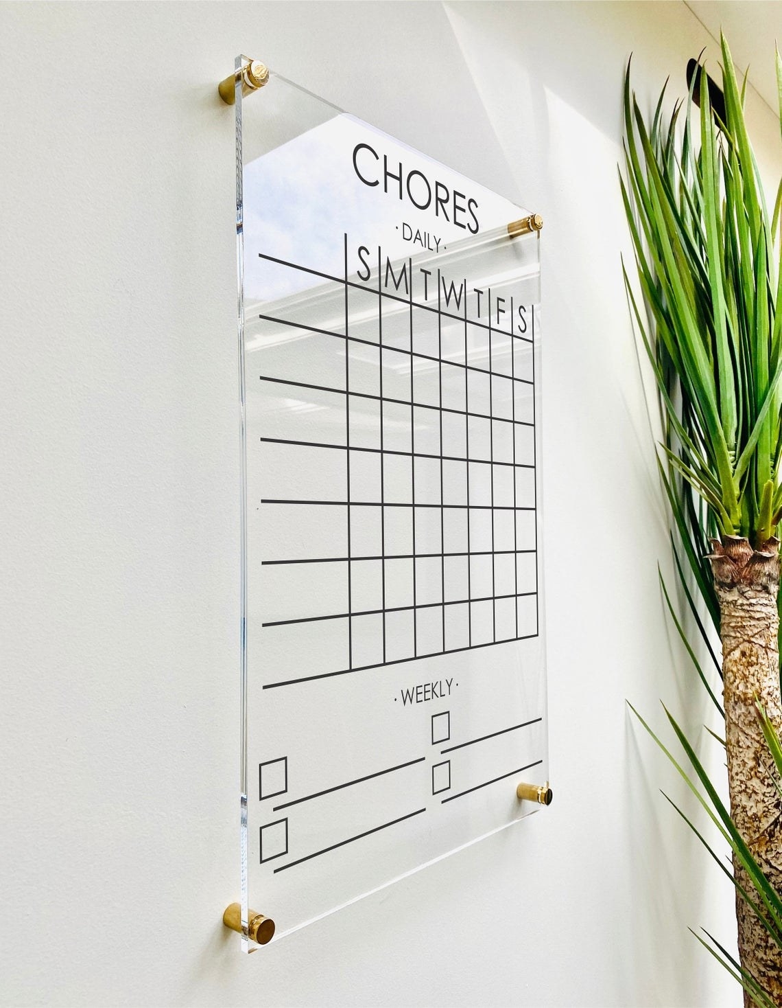 Clear acrylic chore chart mounted on wall with golden attachments on every corner. The chart has spots for daily and weekly chores. 