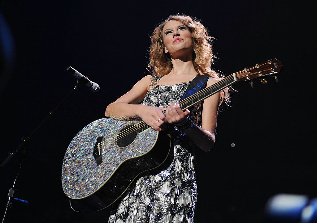 Taylor Swift stands on stage and smiles as she holds a sparkly guitar
