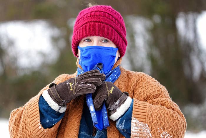 A white woman outdoors wearing a pink beanie, blue cloth mask, brown gloves, and an orange knit sweater