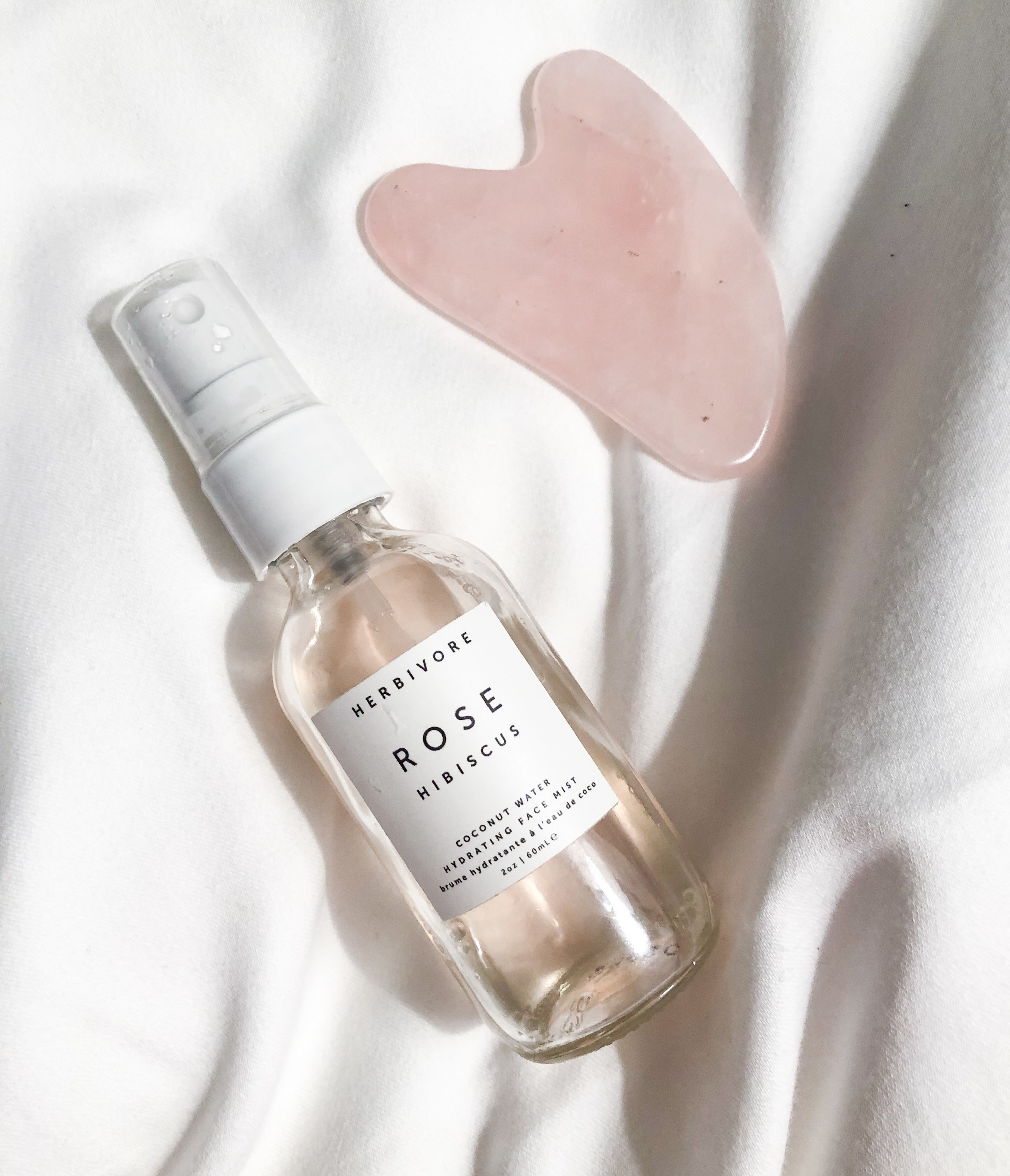 A flatlay of the face mist on clean sheets next to a rose quartz gua sha tool
