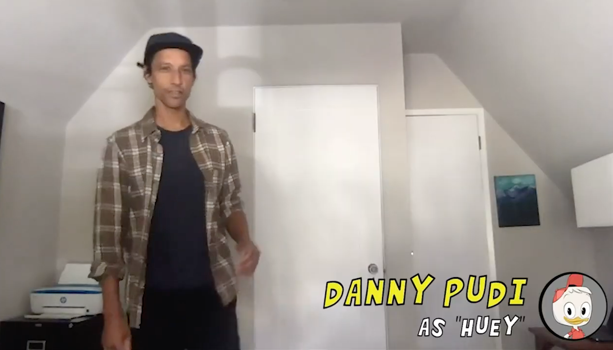 Screen shot of Danny Pudi standing and wearing a flannel shirt and a baseball cap