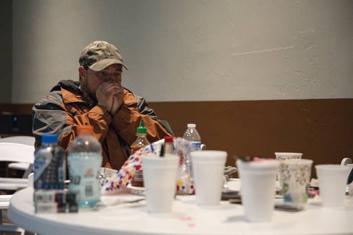 A man sitting indoors wearing a jacket and cap exhales into his hands, cupped over his mouth, in an effort to keep warm