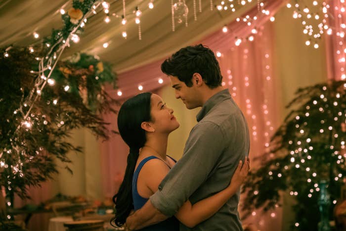 Peter Kavinsky and Lara Jean smile at each other while embracing in a tent lit up with fairy lights