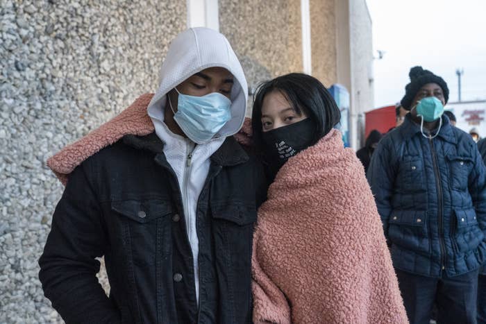 Two people wearing face masks and sharing a blanket around their shoulders