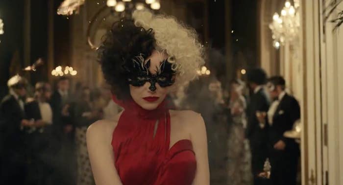 Cruella with two-toned hair and a mask on her face as she attends what appears to be a masquerade