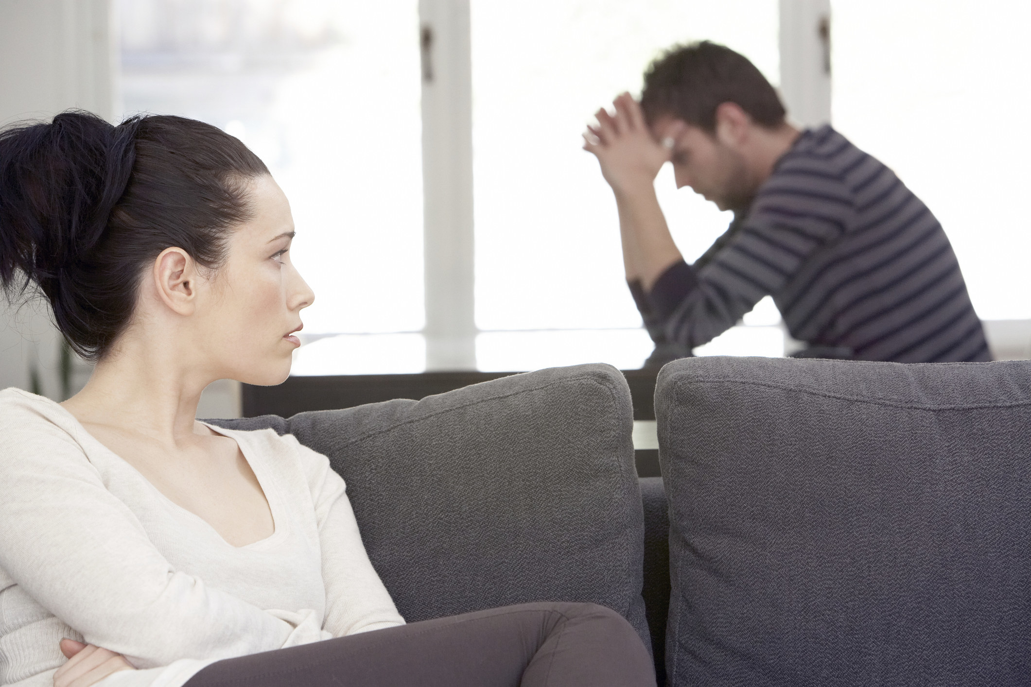 A woman sits on a couch looking at a man behind her who is leaning on his elbows as he contemplates something