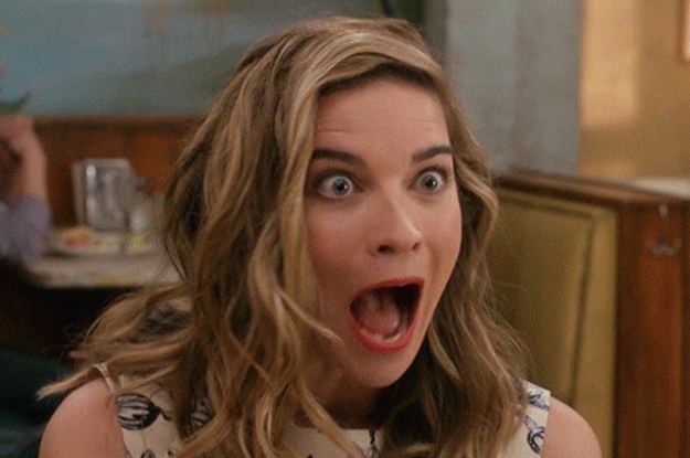 woman with wavy hair sits in a cafe with her mouth and eyes wide open, eyebrows raised