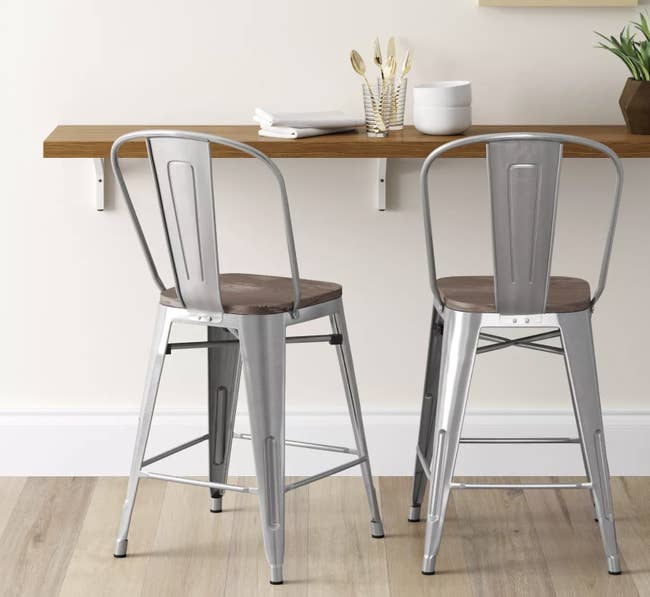 two silver metal bar stools with metal legs, a backrest, and a wooden seat, propped in front of a bar