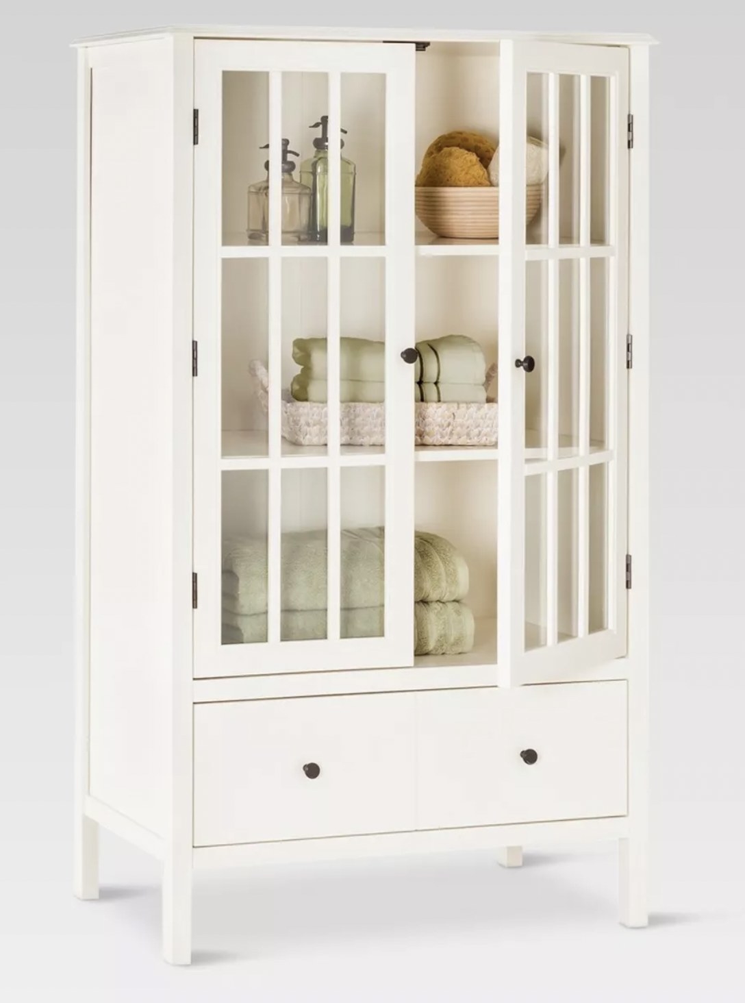 a cream colored tall cabinet with three shelves behind glass doors. Under the shelves are two drawers with black metal hardware