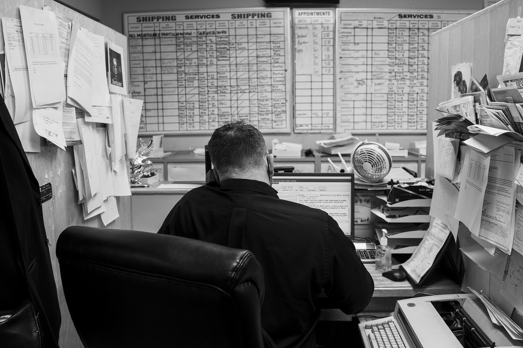 A man seen from behind at a desk covered with papers and a full calendar 