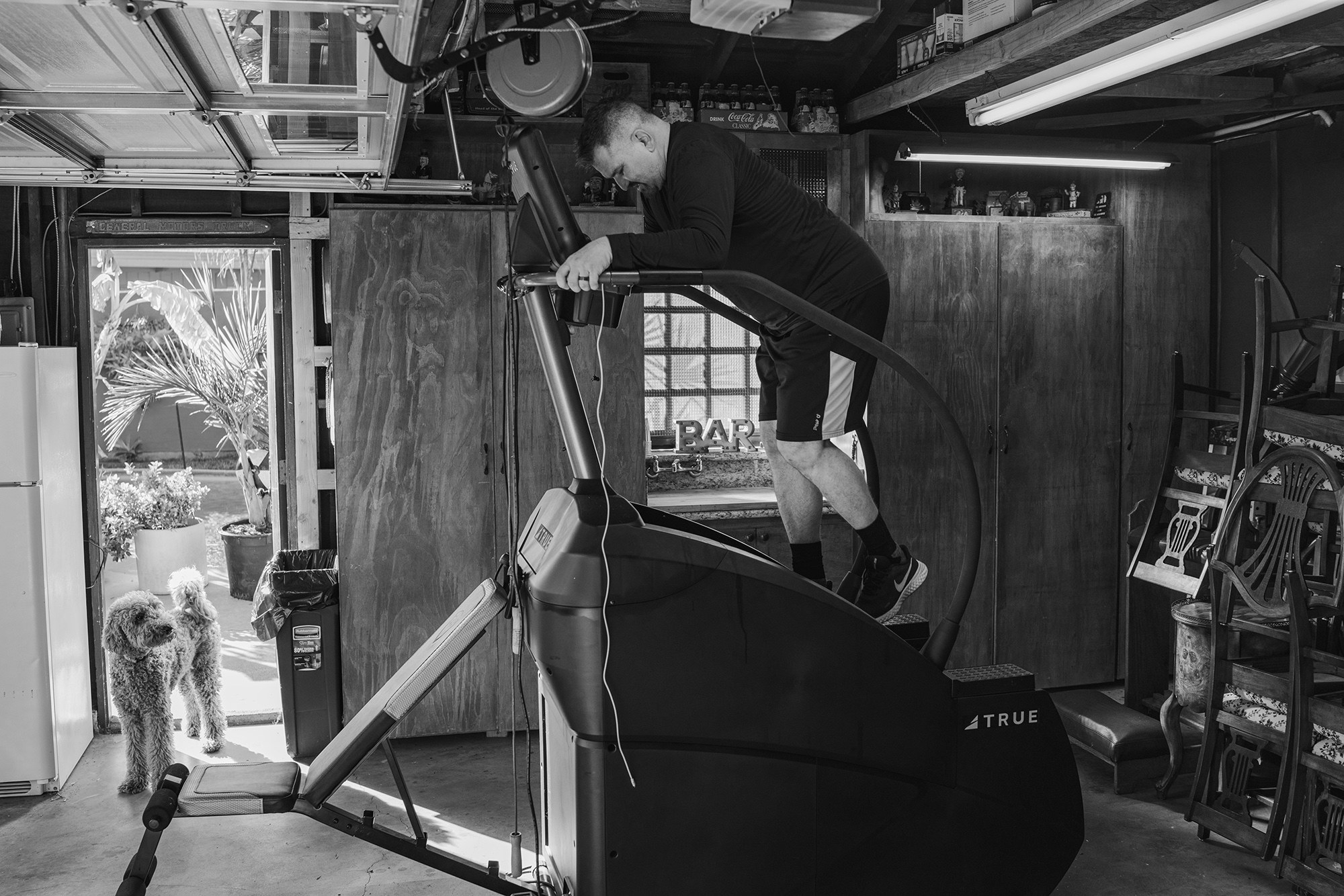 A man on a stairmaster in a garage while a dog watches, a pile of chairs is behind him 