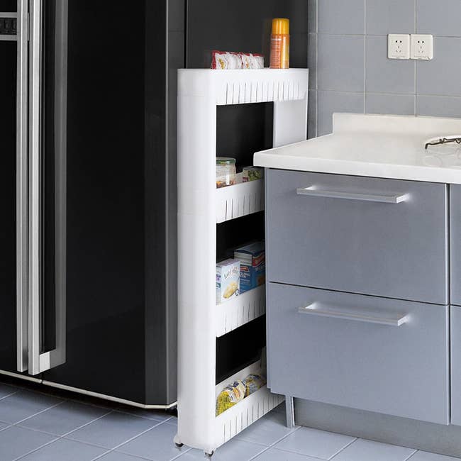 A slim white four-shelved organizer with wheels between a fridge and a counter 