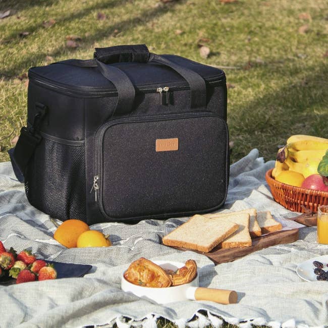 Canvas cooler with handles, crossbody straps, and several pockets on top of a picnic blanket with food around it 