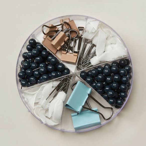 A transparent circular organizer filled with binder clips, push pins, and paper clips