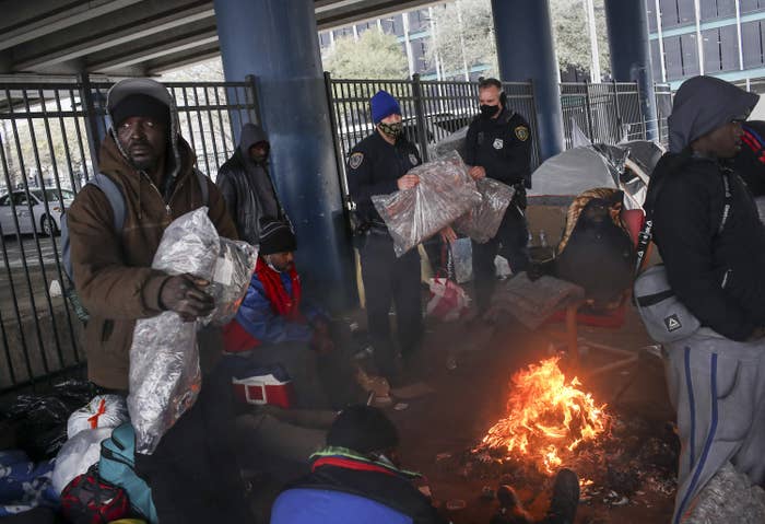 Police wearing face masks carry plastic-wrapped blankets and pass them out among people experiencing homelessness who huddle near a fire