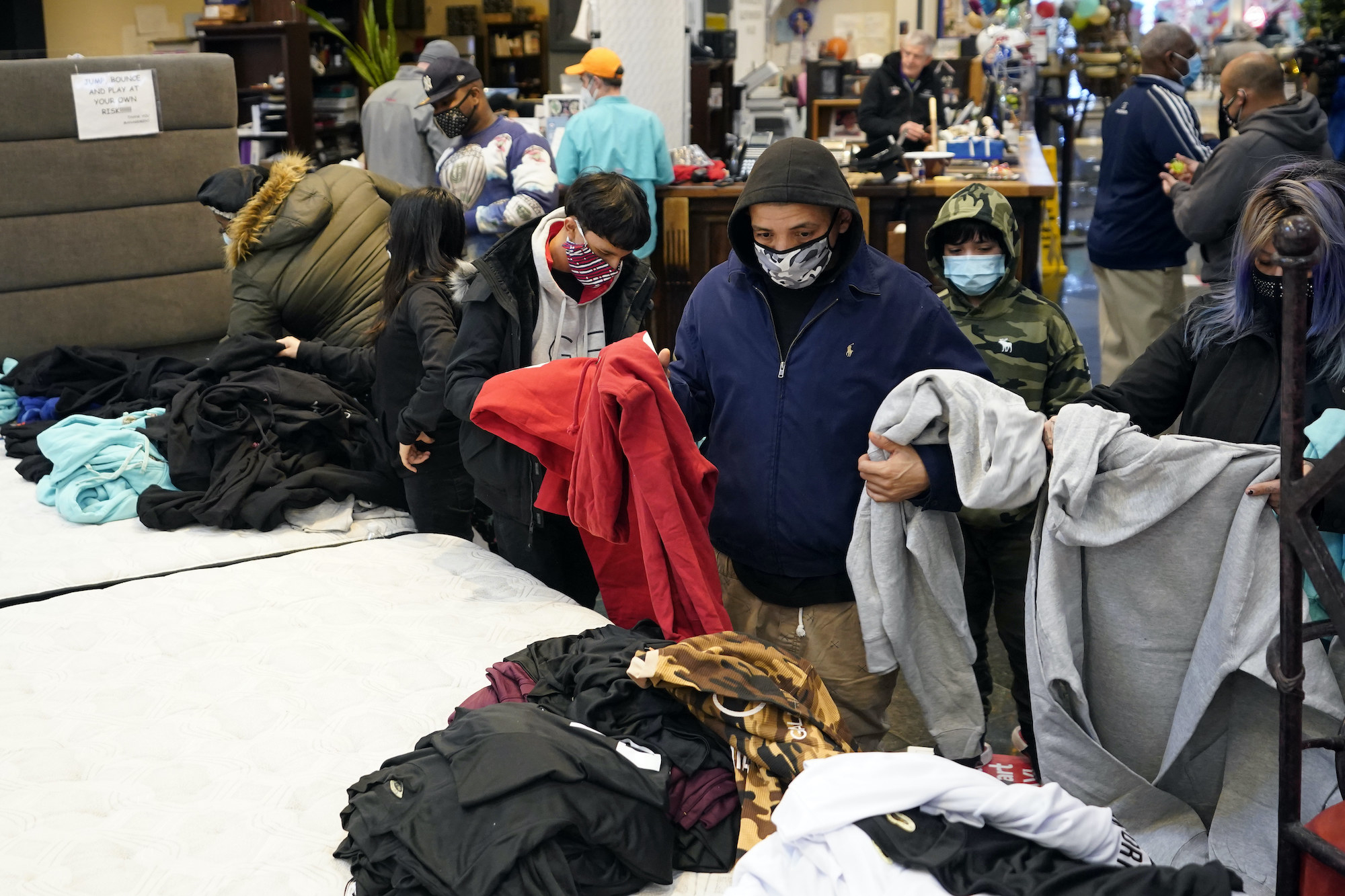 A person wearing a hoodie holds sweatshirts and looks through piles of clothing inside a furniture store