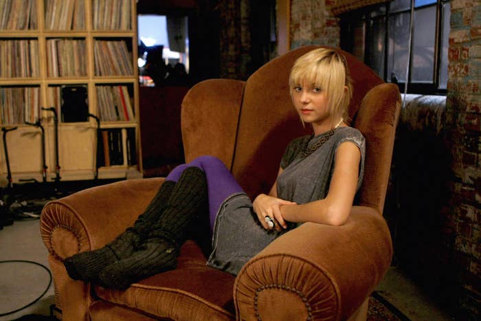 Taylor Momsen sits on a chair in an episode of Gossip Girl wearing a dress, tights, and knee-high socks