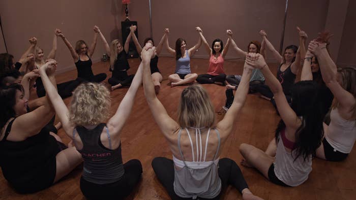 A group of women in athletic wear sit in a circle with hands joined and raised between them