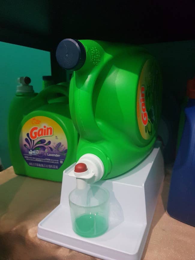 the two-tier white stand holding a sideways detergent bottle on top and a detergent cup below, so the bottle can pour right into it