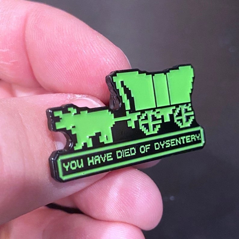 hand holding a pin that looks like the 8-bit style of the Oregon Trail video game with &quot;you have died of dysentery&quot; on it
