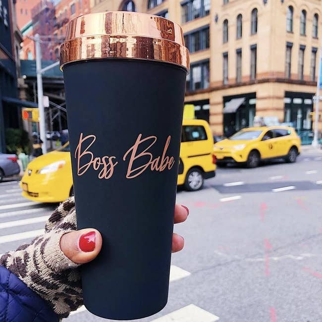 a hand holding the boss babe rose gold matte black travel mug on a busy city street