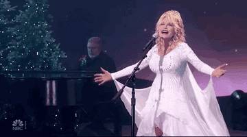 Dolly wearing a dress with batwing sleeves raises her arms as she sings onstage 