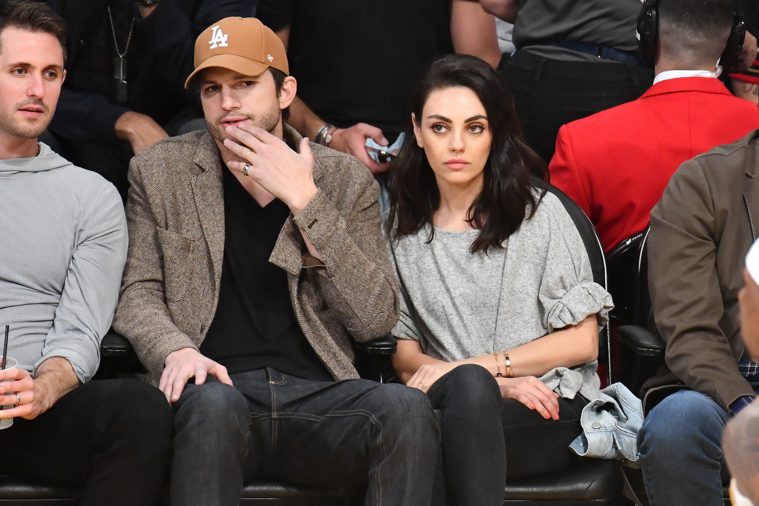 Ashton Kutcher, wearing a Los Angeles Dodgers baseball cap, jacket, v-neck, and jeans sits next to Mila Kunis, wearing a casual shirt and pants, while watching a Los Angeles Lakers Game