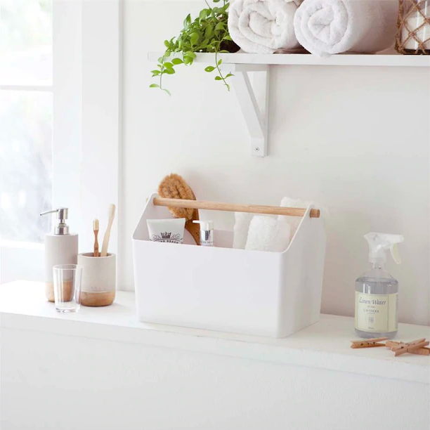 A rectangular basket with wooden handle that's filled with towels soap, and other bathroom items