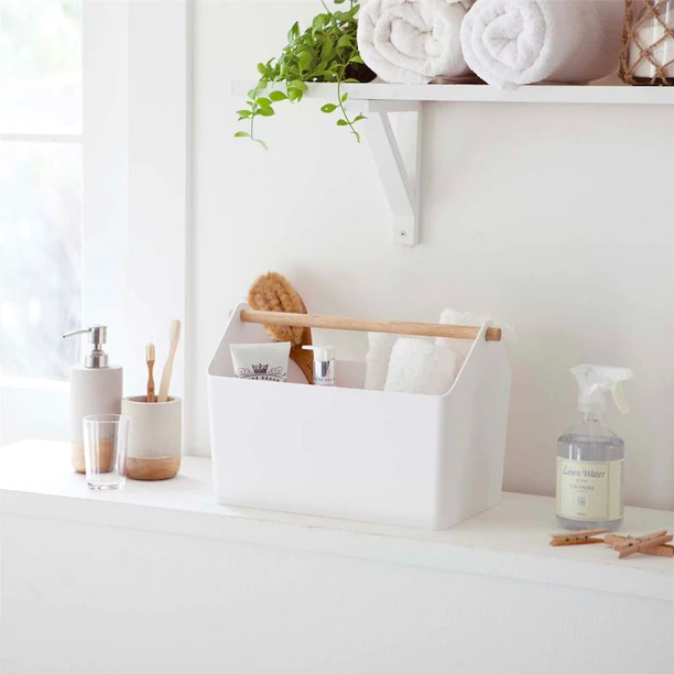 A rectangular basket with wooden handle that&#x27;s filled with towels soap, and other bathroom items