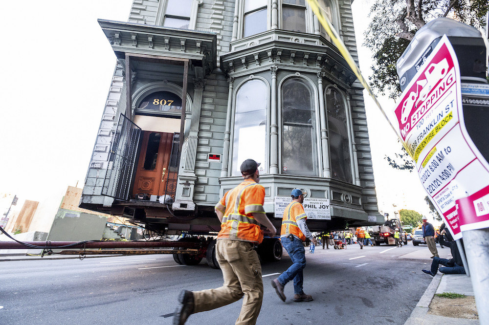 Two workers run behind house on wheels being moved down the street