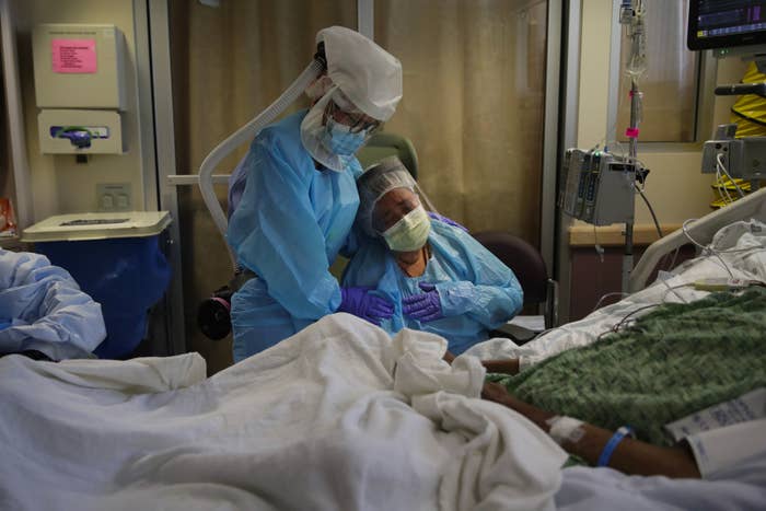 A nurse, wearing a face shield and other protective gear, holds a woman, also wearing PPE, who cries with a gloved hand pressed to her chest while sitting beside a hospital bed