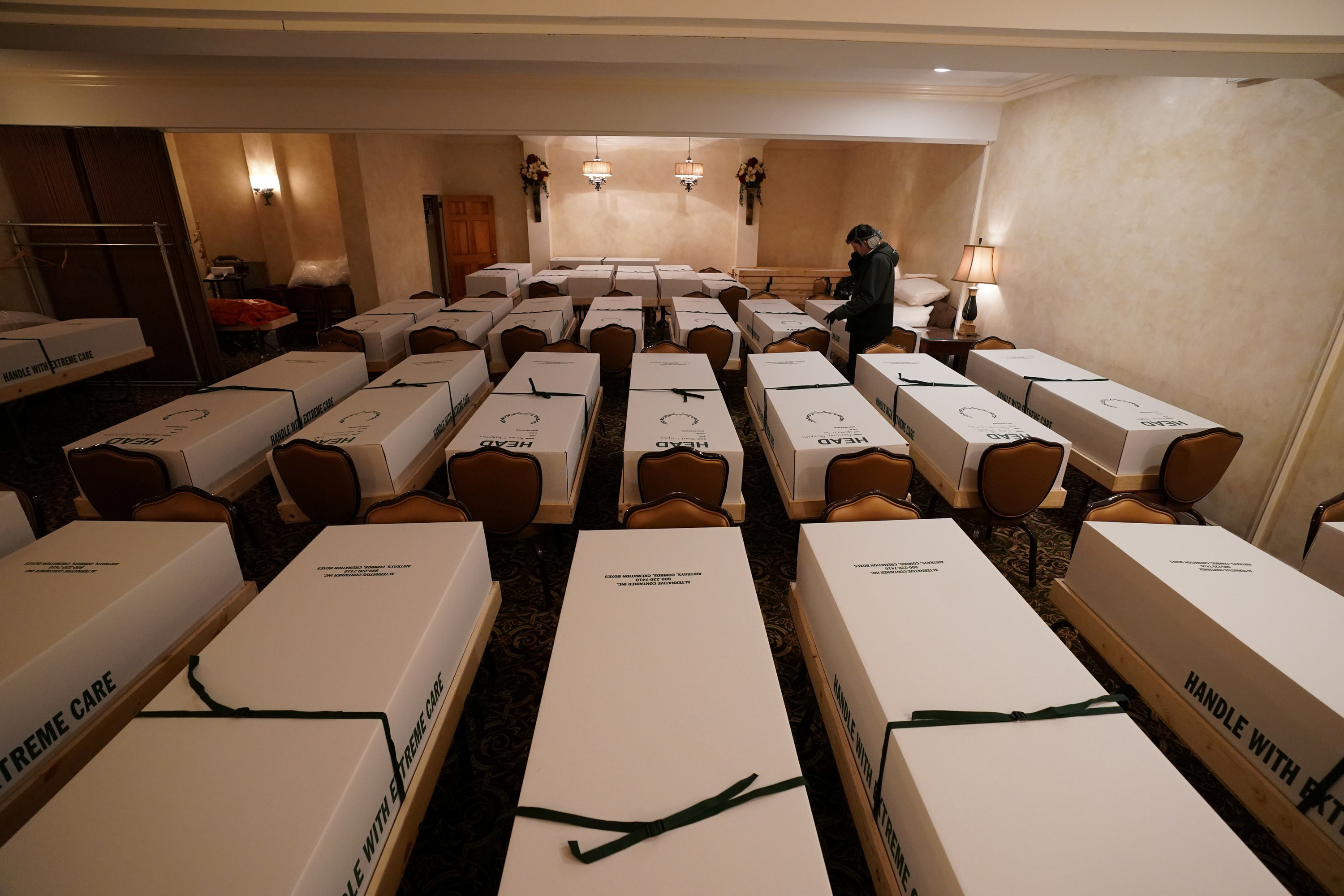 A camera overlooks multiple rows of caskets that rest on chairs inside a funeral home as one man stands in the middle of them