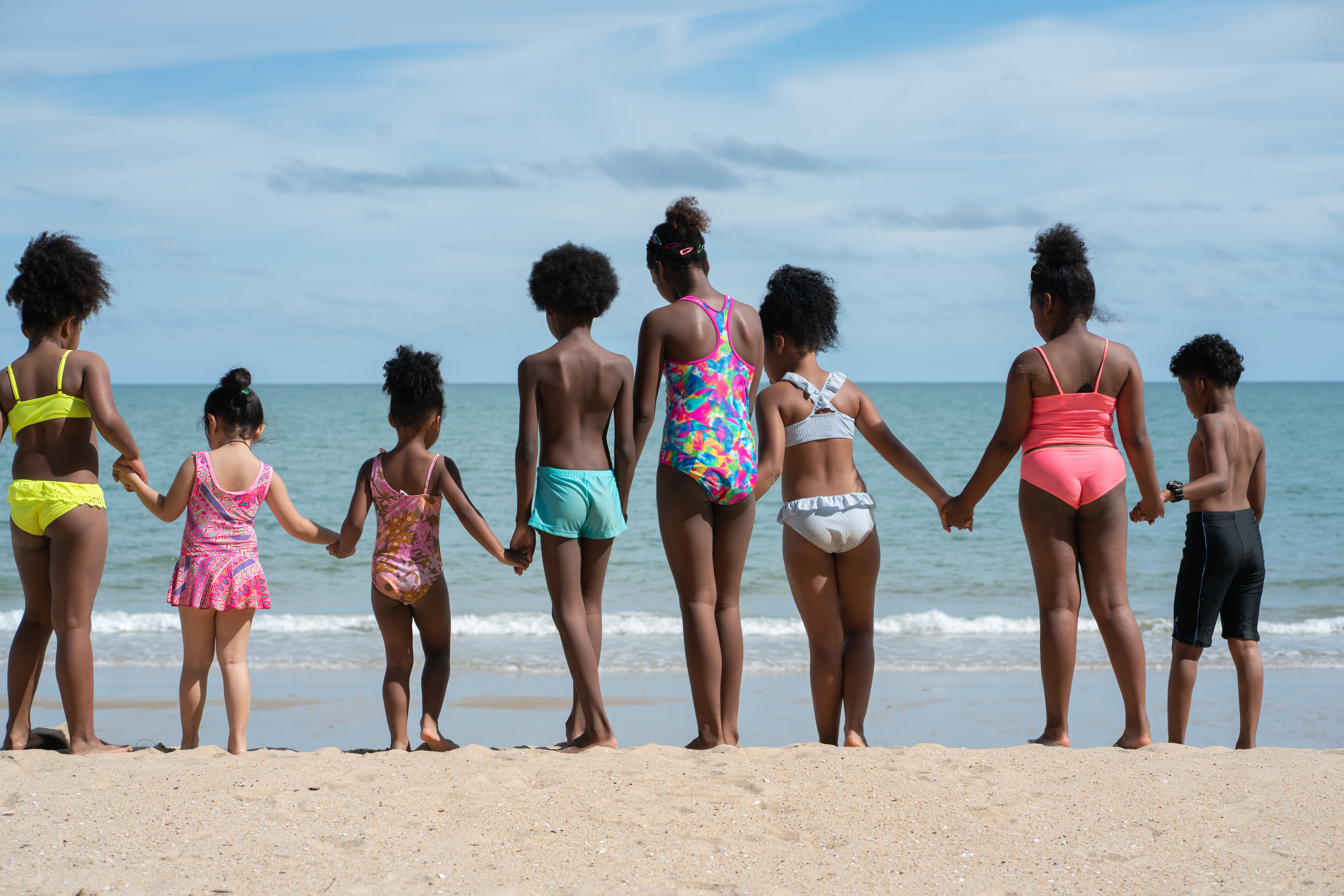 Black children hold hands, wearing swimsuits and standing in a row on the shore of a beach, facing the ocean