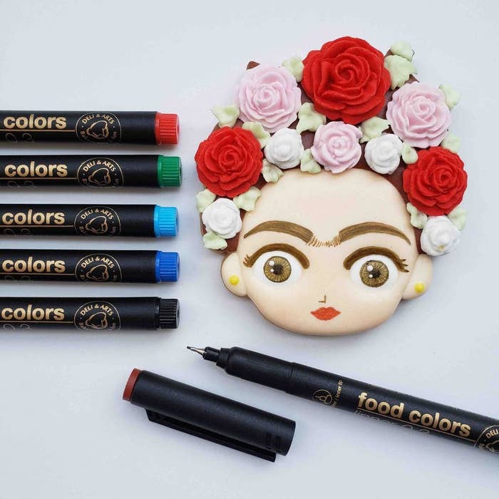 A bunch of markers next to a cookie that looks like Frida Kahlo