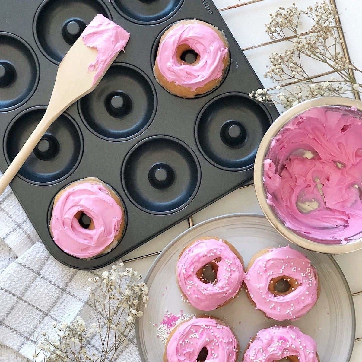 A metal donut pan with two iced donuts beside a plate of more donuts and a bowl with icing