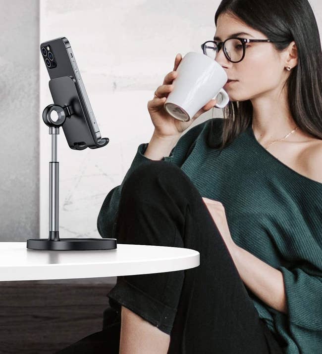 A model watching something on their phone which is propped up to eye level on a black stand on a desk 