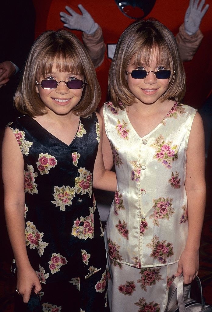 A young Mary-Kate and Ashley wearing matching satin tops and skirts with a floral print and sunglasses