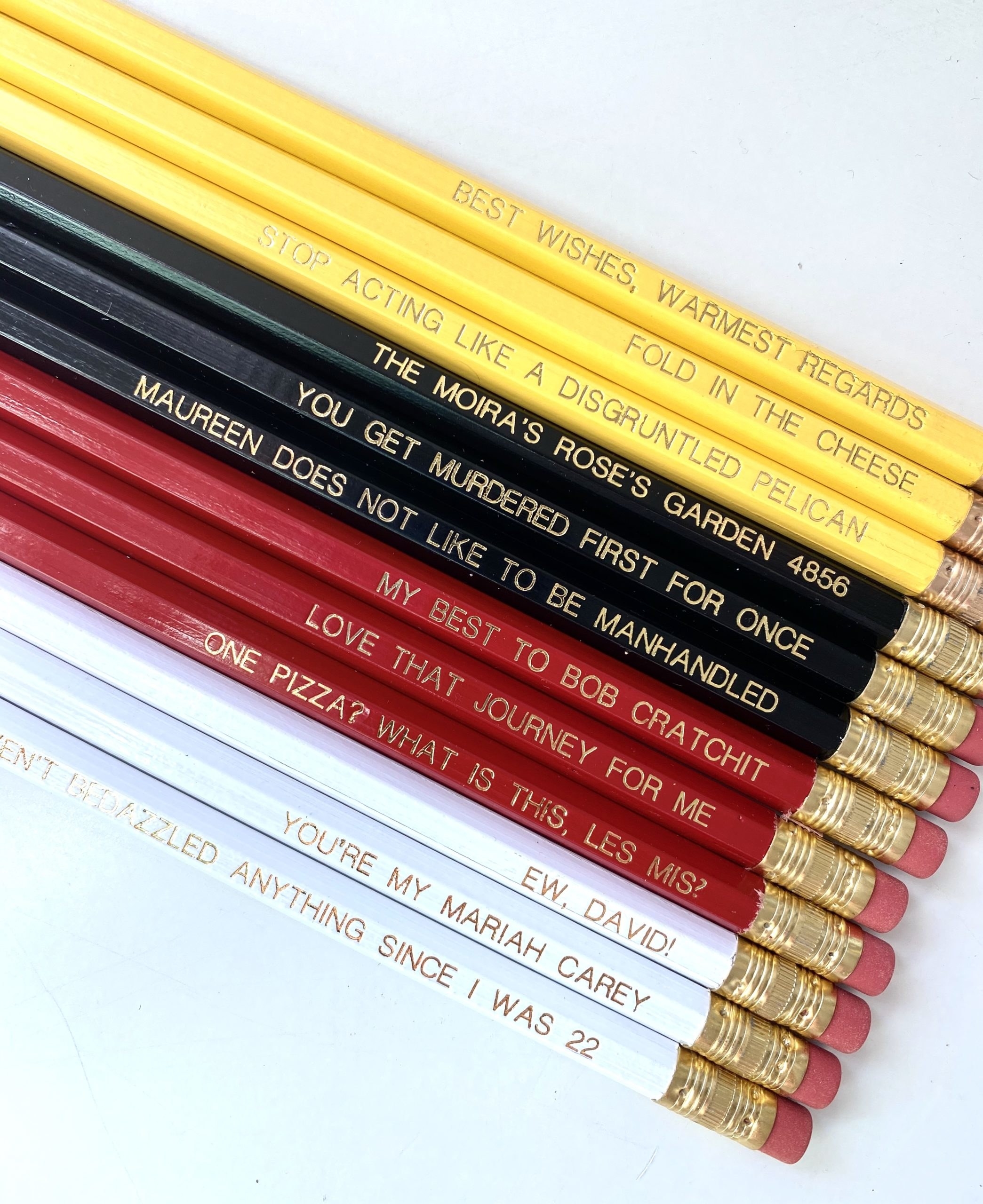 the set, featuring three yellow, three black, three red, and three white pencils each with a different quote in gold text, from classics like "Fold in the cheese" to more obscure gems like "I haven't bedazzled anything since I was 22"
