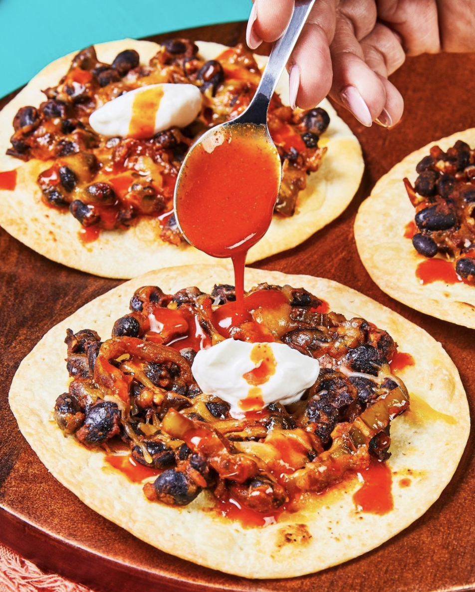 person drizzling hot sauce on Cheesy Black Bean Tostadas
