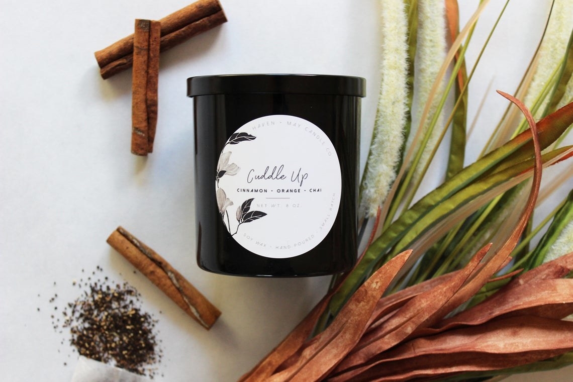 Black jar candle with lid beside cinnamon sticks and plants 